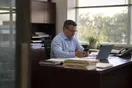 An accountant sits at his desk in a small office checking his phone during a quiet moment in late afternoon, with a stack of folders and a laptop open in front of him, in a natural documentary style.