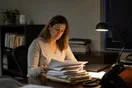 An accountant sits at her desk after hours reviewing a stack of client files beside a quiet phone, in a natural documentary style.