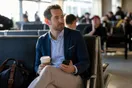 A person in business casual clothing checks a phone while waiting near airport gate seating, photographed in a natural documentary style.