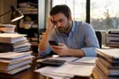 A CPA sits at a desk stacked with folders, checking a slow-loading page on his phone during tax season, in a natural documentary style.