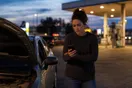 A woman stands beside her car in a gas station parking lot at dusk, checking her phone after the engine started making a strange noise.