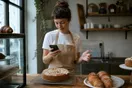 A baker wipes flour from her hands and checks her phone at the counter of a small bakery in the early morning light.
