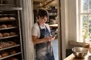 A bakery owner in a flour-dusted apron checks her phone in a sunlit kitchen doorway while a tray of pastries cools nearby, natural documentary light.