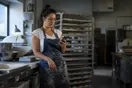 A baker in a flour-dusted apron leans against a stainless prep counter checking a slow-loading page on her phone before dawn, in a natural documentary style.