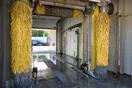 A car wash building on a busy commercial corner with a lit sign and cars queued at the entrance, in a natural documentary style.