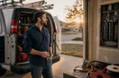 An electrician at the open doors of a freshly equipped work vehicle in early-morning light, keys in hand, in a natural documentary style.