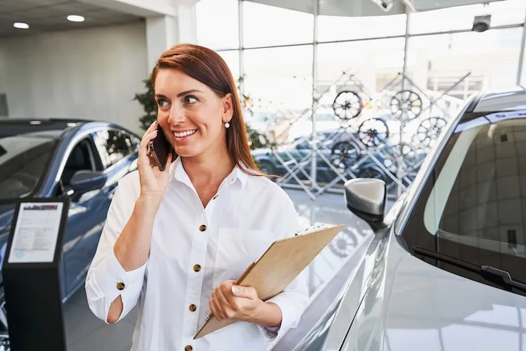 A used-car dealer walking a row of vehicles on a small lot, checking a phone, in a natural documentary style.