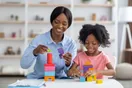 A childcare provider sitting on the floor with toddlers and stacking blocks in a bright playroom, in a natural documentary style.