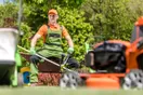 A landscaper unloading a push mower and string trimmer from an open utility trailer on a suburban street, in a natural documentary style.