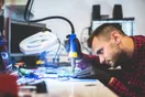 A phone repair technician at a workbench opening a smartphone with a spudger under a task light, in a natural documentary style.