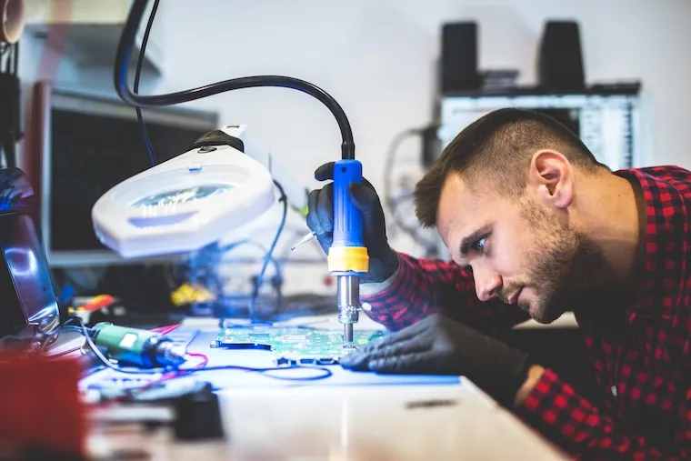 A phone repair technician at a workbench opening a smartphone with a spudger under a task light, in a natural documentary style.