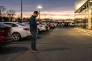 A car buyer sits in a parking lot outside a dealership at dusk, scrolling on their phone before walking in, with rows of vehicles softly lit in the background, in a natural documentary style.