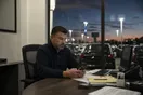 A car dealership owner checks a quiet phone at his desk overlooking a lit lot of cars at dusk, in a natural documentary style.