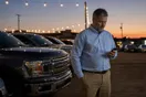 A car dealership owner standing on the lot at dusk, checking his phone beside a row of parked vehicles under the lot lights.