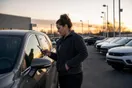 A person stands in a car dealership lot at dusk holding a phone up next to a vehicle window sticker, photographed in a natural documentary style.