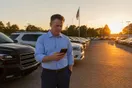 A car dealership general manager checks a slow-loading vehicle listing on his phone while standing among rows of parked cars on the lot at sunset, documentary style.
