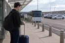 A traveler stands at an airport curb with a rolling suitcase, checking a phone while a shuttle idles nearby and a row of rental cars waits in the distance, in a natural documentary style.