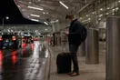 A traveler stands at an airport curb at night with a rolling suitcase, looking at a phone, photographed in a natural documentary style.