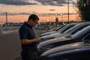 A rental car lot attendant checks his phone near a row of parked vehicles as evening falls near an airport access road.