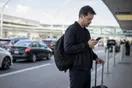 A traveler stands near an airport curb checking his phone with a rolling suitcase beside him, waiting for a page to load, in natural documentary light.