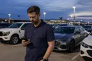 A car rental operator checks a slow-loading booking page on his phone near a small fleet of parked vehicles at an airport lot, documentary style.