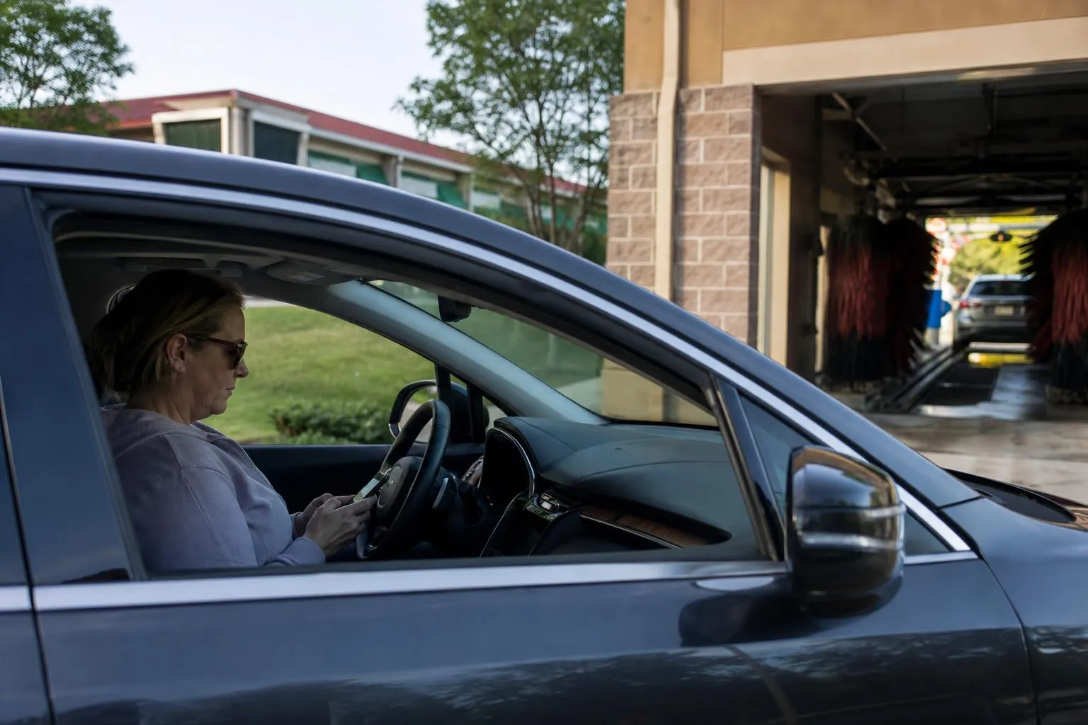 A driver sits in a parked car at a strip mall checking their phone before pulling into a car wash entrance, with a wash tunnel visible in soft afternoon light, in a natural documentary style.