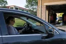 A driver sits in a parked car at a strip mall checking their phone before pulling into a car wash entrance, with a wash tunnel visible in soft afternoon light, in a natural documentary style.