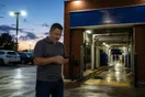 A car wash owner checks a phone near the entrance tunnel of his wash at dusk with the lot lights coming on, in a natural documentary style.
