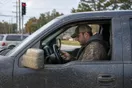 A driver sits behind the wheel of a muddy pickup at a red light holding a phone, photographed in a natural documentary style.