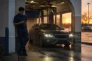 A car wash attendant leans against the entrance archway checking his phone as a customer's car exits the tunnel at dusk.