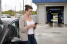 A driver stands beside her car in a wash bay parking lot checking her phone, waiting for a page to load, in natural documentary light.