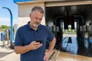 A car wash owner checks a slow-loading membership signup page on his phone near the tunnel entrance on a bright afternoon, documentary style.