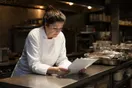 A caterer stands beside her prep table late at night reviewing an event tasting order sheet, in a natural documentary style.