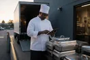 A caterer in chef whites checking a tablet next to stacked chafing dishes in a commercial kitchen loading dock at dawn.