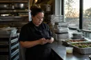 A caterer stands in a stainless steel kitchen scrolling her phone during a lull between prep and a wedding delivery, early evening light through the window.