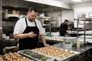 A caterer in a chef coat glances at a phone in a stainless-steel prep kitchen while trays of hors d'oeuvres sit ready, natural documentary light.