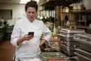 A caterer in a chef coat checks a slow-loading page on her phone beside stacked chafing dishes in a commercial kitchen, in a natural documentary style.