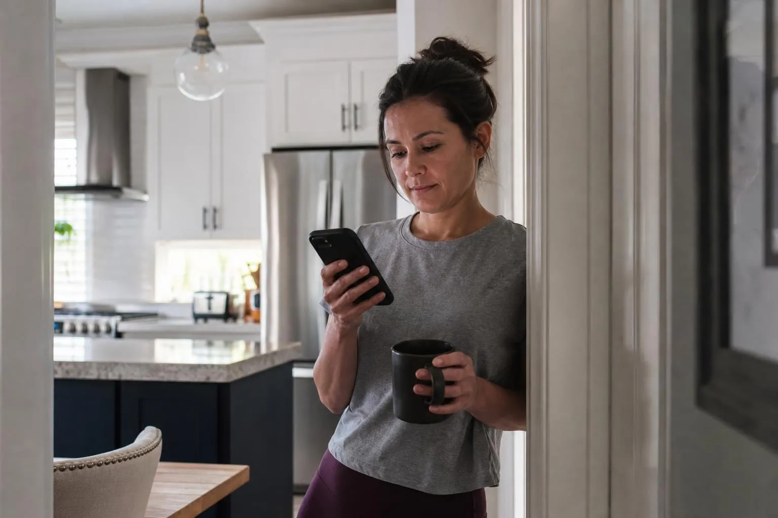 A woman stands in her kitchen doorway on a Sunday morning, phone in hand, comparing cleaning company websites before guests arrive.
