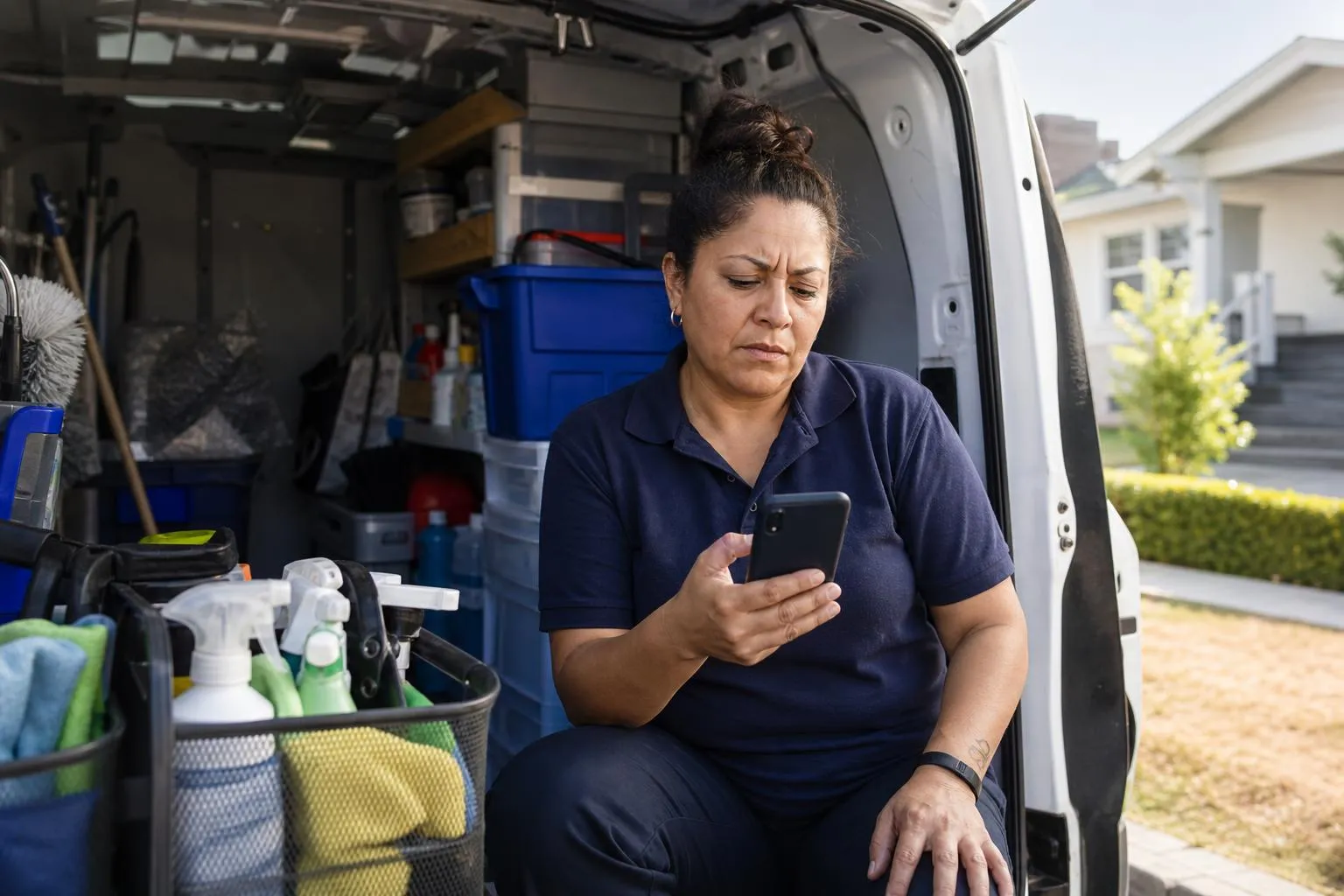 A cleaning company owner sits in her supply van after a job, scrolling a slow-loading quote page on her phone with cleaning caddies stacked in the back, in a natural documentary style.