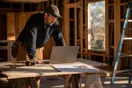 A general contractor stands at a laptop in a half-finished kitchen remodel reviewing project photos in late afternoon light, in a natural documentary style.
