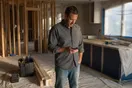 A general contractor stands in an unfinished kitchen remodel scrolling his phone, drop cloths and exposed studs around him in late afternoon light.