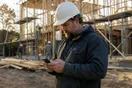 A general contractor checks his phone on a job site with framing lumber and scaffolding behind him, waiting for a page to load, in natural documentary light.