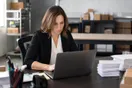 An office manager sits at a desk comparing courier company websites on a laptop with a stack of shipping labels nearby, mid-afternoon office light, documentary style.