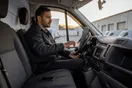 A courier company owner sitting in the driver's seat of a parked cargo van at dusk, phone on the seat, checking a laptop resting on the steering wheel.