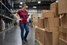 A logistics coordinator stands on a warehouse floor near stacked packages, checking a phone for a same-day courier's website, photographed in a natural documentary style.