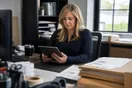 An office manager reviews courier company websites on a tablet at her desk during a short break, a stack of shipping labels beside her keyboard.