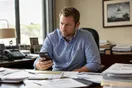 A person in an office reviews a phone screen at a desk cluttered with paperwork, waiting for a courier company's website to load.