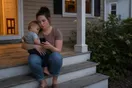 A mother sits on a porch step scrolling her phone with a toddler on her hip, comparing daycare websites in the early evening light, documentary style.