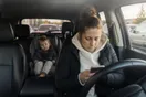A mother sits in a parked car outside a preschool, scrolling a phone with a toddler buckled into a car seat behind her, photographed in a natural documentary style.