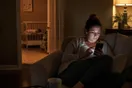 A mother sits on her couch late at night, phone lit up in a dim living room, scrolling through daycare websites while her toddler sleeps nearby.